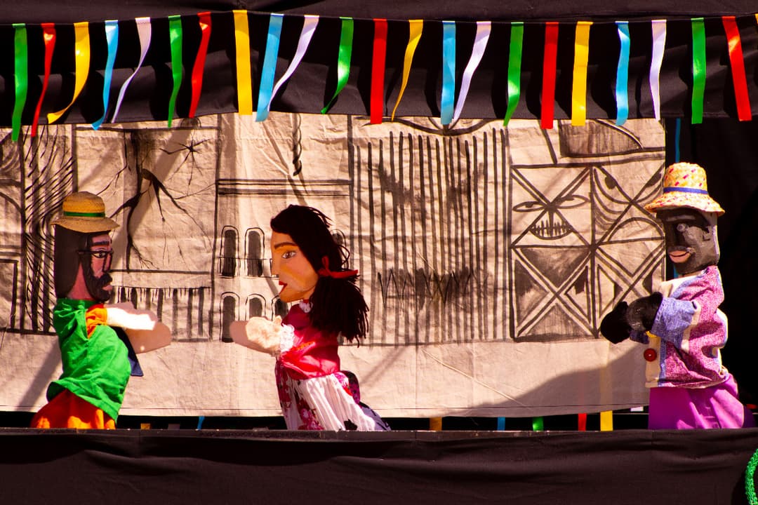 Três bonecos de teatro em cena com fundo abstrato e detalhes geométricos.