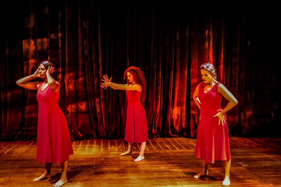 Três mulheres em vestidos vermelhos, dançando em palco com fundo escuro e iluminação cênica.