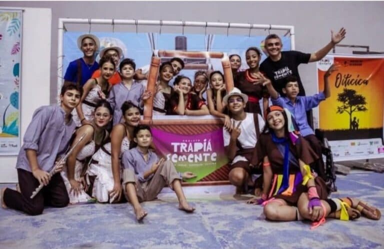 Grupo posando para foto em frente a cartaz com o texto "TRAPIÁ SEMENTE".
