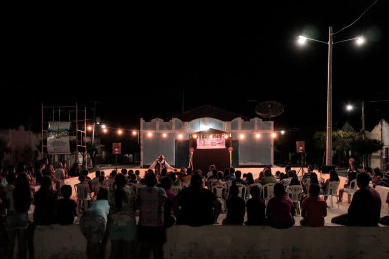 Apresentação ao ar livre à noite, com plateia em cadeiras plásticas e palco iluminado por luzes. Um artista se destaca no centro do palco.