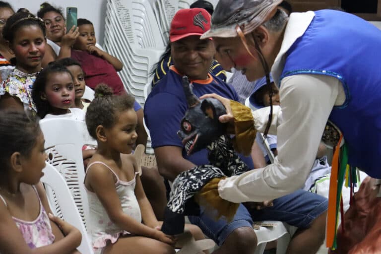Um artista vestido com traje colorido e máscara interage com uma criança sentada em cadeira plástica, enquanto outras crianças e adultos observam atentamente.