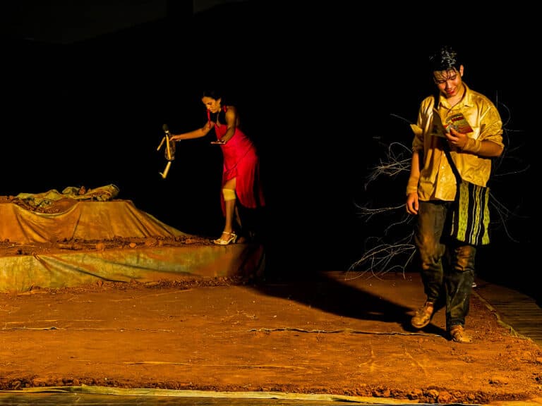 Atriz em vestido vermelho segura boneca, enquanto ator lê livro.