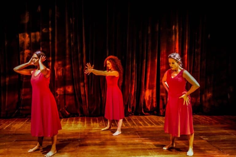 Três mulheres em vestidos vermelhos, dançando em palco com fundo escuro e iluminação cênica.