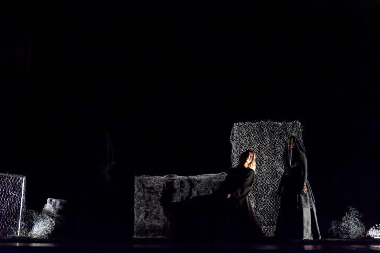 Em um palco escuro, três figuras vestidas de preto estão em cena. Uma mulher de cabelos brancos senta-se, amparada por outra. Ao fundo, painéis de rede cinza compõem o cenário.